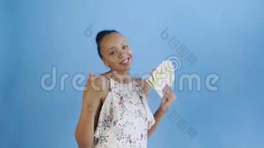 一位非洲裔美国妇女正在演播室里用金钱跳舞，并在蓝色背景上竖起大拇指。带花的白色连衣裙
